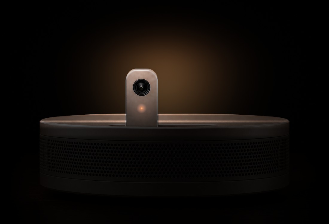 A Nodi home device: a low, round speaker in warm silver, its small camera raised from the top with a coral light under the lens